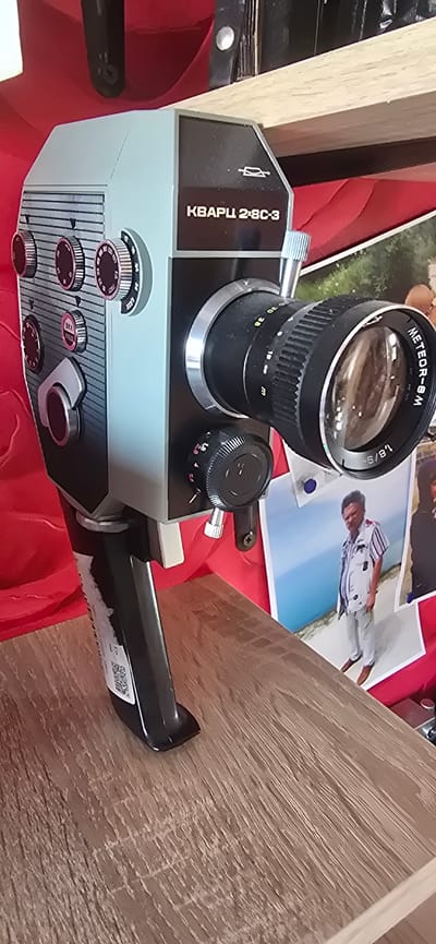 The image shows a close-up, slightly angled view of a vintage, light blue and black film camera with a large, silver and black lens. The camera has a black handle extending to the right. It rests on a red surface, possibly fabric, with several photographs scattered around it. One photograph in the background shows a person in a white and striped outfit, lying down on a blue surface. The camera has several dials and buttons on its body. The lens is labeled "METEOR-8M 1,8/9-36". The body of the camera also has text that appears to be in Cyrillic, reading "КВАРЦ 2-8С-3". The setting appears to be indoors, possibly a home or a shop display, given the presence of the camera and photographs. There are also hints of wooden surfaces in the background, suggesting furniture or shelving. The overall impression is that of an aged piece of equipment, possibly being showcased or used for display. The context provided indicates the location is Gostagayevskaya, Russia. No specific time of day, weather, or discernible emotions are evident.