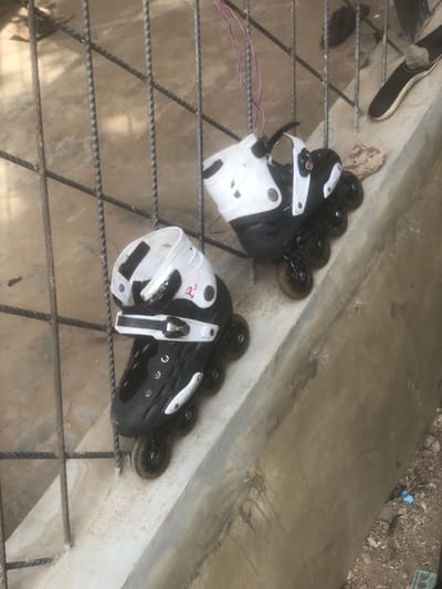 Location: Kufena, Nigeria.
The scene presents an outdoor environment featuring two inline roller skates resting on a sloped, unrefined concrete structure. A grid of vertical and horizontal metal bars, likely rebar, occupies the foreground. Both roller skates are primarily black with white plastic cuffs, adjustable straps, and four dark wheels each. The lower skate exhibits a red marking on its white cuff. A thin purple cord hangs vertically near the upper roller skate. To the upper right of the concrete structure, a single black shoe and a small light-colored rock are visible. The ground visible through the metal grate and to the right of the concrete consists of light brown dirt and darker soil mixed with minor debris. Natural daylight illuminates the scene.