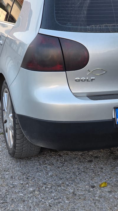 The image shows the rear-left side of a silver Volkswagen Golf. The car has tinted taillights and a chrome "GOLF" emblem with a fish symbol above it. The car is parked on a gravel surface, possibly a street or parking lot in the city of Bania, Bulgaria, given the presence of a license plate that appears to be Bulgarian. There is a small yellow leaf on the ground near the rear bumper. The environment appears to be outdoors, with some building reflections visible on the car's body. The image is a close-up of the car's rear, focusing on the taillight, emblem, and bumper.