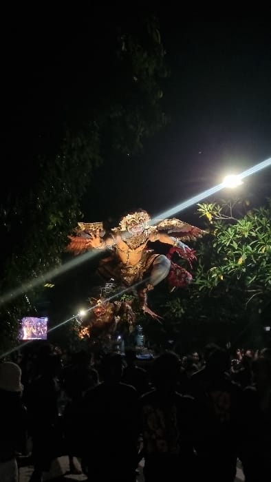 A large, illuminated Ogoh-ogoh effigy dominates the center of a nighttime scene in Pecatu, Indonesia. The effigy depicts a multi-limbed, fierce-faced humanoid figure with prominent golden adornments and red accents, caught in a dynamic, crouching posture above a smaller, darker creature. Bright artificial light sources cast a strong glare and horizontal lens flares across the upper-mid section of the image, illuminating the effigy against the dark sky. Dense, silhouetted crowds of individuals stand in the foreground, facing towards the effigy. Green foliage from trees is visible on the left and right sides of the main subject. A small illuminated screen displaying multiple human figures is situated in the lower-left background.