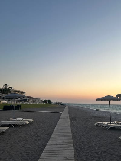 This media file depicts a beach scene at sunset in Rhodes, Greece. The image is oriented vertically, with the sky occupying the left half and the beach and resort on the right. The sky displays a gradient of colors, transitioning from a pale blue at the top to soft orange and yellow hues near the horizon, indicating it is dusk.
The beach itself is covered in dark sand and pebbles. A wooden boardwalk runs along the edge of the beach, providing a path. Several empty white lounge chairs with umbrellas are scattered across the sand, suggesting a resort area. In the background, there are modern hotel buildings with white facades and balconies, surrounded by green lawn areas. Palm trees and other Mediterranean vegetation are visible, adding to the tropical resort feel.
The sea is calm and appears in shades of blue-grey. There is a small, distant structure or building visible on the horizon near the sea. The overall atmosphere is serene and peaceful, characteristic of a quiet evening by the sea. No people are prominently visible in the frame, though the presence of lounge chairs and resort buildings implies human activity and vacationing. No visible text is present in the image.