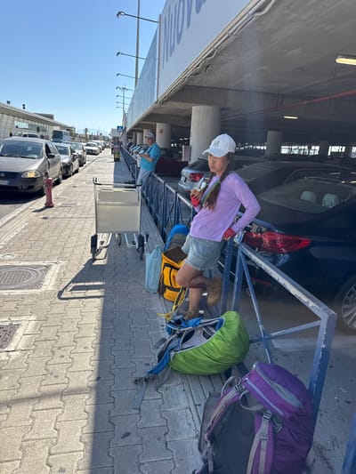 The image depicts a woman standing near a parking lot in Cinisi, Italy. She is wearing a white cap, a long-sleeved lavender shirt, denim shorts, and brown hiking boots. She also has red gloves on her hands. A large purple and black backpack, along with a green-covered backpack, are situated on a blue metal structure behind her. There are several suitcases and a yellow bag with blue straps next to her on the ground. In the background, a row of cars are parked alongside the sidewalk, and a fire hydrant is visible near the curb. The ground is paved with interlocking grey bricks. Overhead, there is a parking structure with cars parked inside. A sign is also visible on the left side of the image.