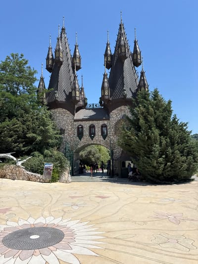 The image depicts a fantastical castle structure with multiple spires and a stone facade, set against a clear blue sky. The castle is surrounded by lush green trees and foliage. In the foreground, a textured pathway is visible, decorated with large, stylized floral patterns, resembling sunflowers and other blossoms. Several people are seen near the entrance of the castle, some with strollers, suggesting it might be a tourist attraction or park. The lighting suggests it is daytime, likely sunny. The overall scene evokes a sense of wonder and enchantment, possibly a fairy tale-themed location. There is a sign visible on the left, partially obscured by foliage, which appears to be a poster or information board, but the text is illegible. The architectural style of the castle, with its pointed roofs and ornate details, points towards a whimsical or gothic revival design. The presence of people suggests an ongoing public event or the castle being open to visitors.