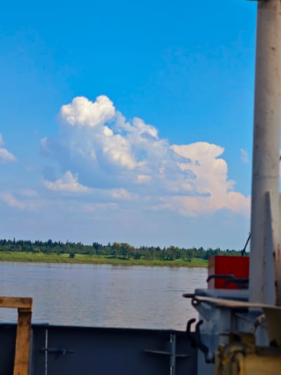 The image depicts a view from a vessel on a body of water, likely a river, with the distant shoreline visible on the horizon. The sky is bright blue with large, fluffy white clouds. The water appears calm, reflecting the sky. The foreground shows parts of the vessel, including a metal railing and some equipment, with a wooden structure on the left. The shoreline is lined with a dense forest of green trees. The overall scene suggests a sunny day in a rural or natural setting. Given the context provided, this scene is likely located in or near Усть-Кемь, Russia, a town situated on the Kema River.