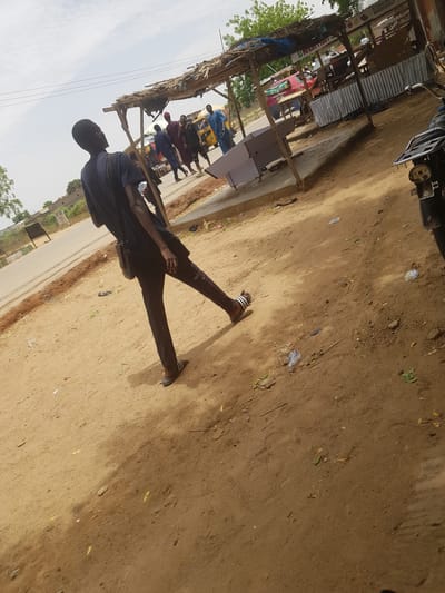 A medium shot captures a man in Dabawa, Nigeria, from a low angle while he is walking on a sandy, dirt path. The man is wearing dark clothing and sandals and appears to be carrying a bag or satchel. He is walking away from the camera along the path. The path extends into the background, where other people are visible near a series of roadside stalls with corrugated metal roofs, a common sight in Dabawa. The environment consists of dirt, foliage, and makeshift structures along the road. The presence of people near the stalls suggests a market or commercial area.
