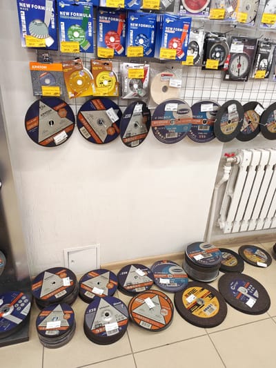The image displays a retail display of cutting discs, primarily for angle grinders, arranged on tiered shelving. The products are predominantly from the "NEW FORMULA" and "КРАТОН" brands, with some "Tsunami" and "GRAND" branded items also visible. The discs vary in size and type, including turbo-segment, ultra-thin, and general-purpose cutting discs, indicated by labels with specifications like diameter (e.g., 125 mm, 230 mm), thickness, and bore size. Many discs have prominent text in Russian, detailing their intended use (e.g., "КРУГ ОТРЕЗНОЙ" - cutting disc, "ПРОФИ" - professional, "СТАЛЬ STEEL" - steel), material compatibility, and safety information. Prices are also visible on many items, with some in the range of 470-00 to 1099-00 rubles.

In the background, a white radiator with red valve handles and plumbing is mounted on a textured, light-colored wall. A small, square white access panel is also visible on the wall. The lower part of the image shows more cutting discs stacked on the floor, some in their original packaging. The lighting appears to be artificial, suggesting an indoor retail environment, likely a hardware store or a specialized tool shop. The overall impression is one of a well-stocked display of tools ready for sale. No people or specific events are depicted.