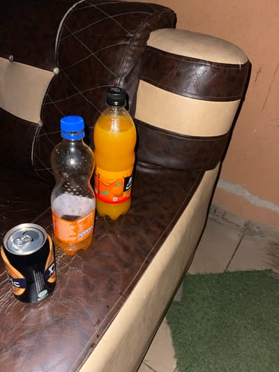 A leather sofa with brown and beige cushions is partially visible. The sofa's arm is adorned with bottles and a can. A partially full orange juice bottle sits near the top of the armrest, below that is a nearly empty soda bottle with a blue cap. A black and gold can lies near the edge. The background reveals a wall, tiles, and artificial green grass.