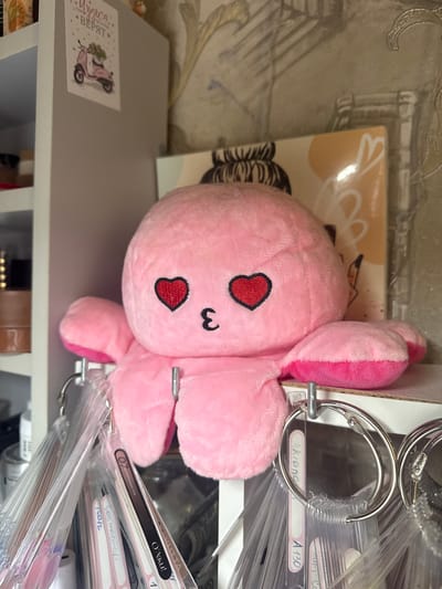 A pink plush octopus toy, featuring embroidered heart-shaped eyes and a "w"-shaped mouth, is prominently displayed, hanging by its tentacles from a silver metal binder ring. The toy is positioned on a light grey shelf or tabletop, appearing to gaze towards the viewer. Behind the octopus, a light-colored box or book cover with abstract white designs is visible.
To the right of the octopus, a collection of transparent plastic swatches, likely for nail polish or paint samples, are neatly organized on several metal binder rings. Many of these swatches bear labels with handwritten or printed text. Visible labels include "Akiano A 130", "MUTTA", "О'Лаки!", "Cosmoprofi", "21021", "Стамм", "B", "10", "3", and "07".
On the grey surface to the left, a small card or sticker depicts a pink scooter adorned with flowers, next to text that reads "Lieca.." and "ВЕРЯТ" (meaning "BELIEVE"). The background features a patterned wallpaper with subtle decorative motifs. Below the shelf, parts of other objects, including a brown item, are partially visible. The scene appears to be an indoor setting, likely a home or hobby space in Novoe Devyatkino, Russia, indicated by the Cyrillic text and location context. No specific activity is taking place; the image captures a static arrangement of items.
