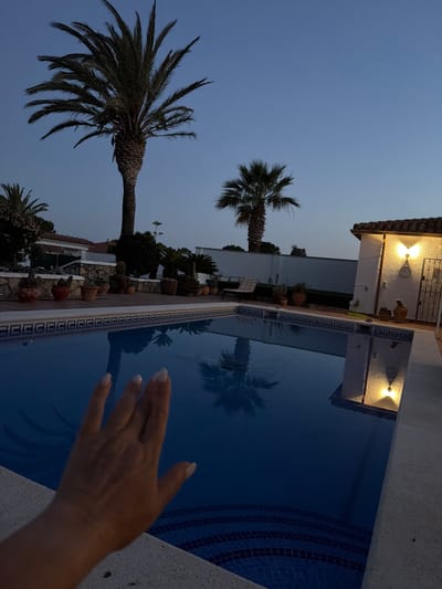 The image captures a twilight scene at a swimming pool in Mont-roig del Camp, Spain. The sky is a gradient of blue. Two palm trees are visible, one in the foreground to the left, and another further back. A white building with lights is in the background, with a structure containing a shower on the right. Numerous plants and pots are arranged along the side of the pool, including cacti and flowering plants. The water in the pool reflects the palm trees and building. A person's hand is partially visible in the foreground on the right side of the image.