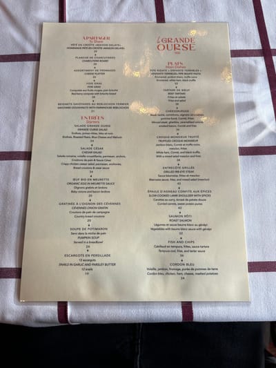 This media file features a restaurant menu, titled "LA GRANDE OURSE 1936," lying diagonally on a white tablecloth adorned with parallel maroon stripes. The setting appears to be indoors, likely a restaurant in Val-d'Isère, France. Glimpses of dark clothing, possibly belonging to a diner, are visible in the top and bottom right corners of the frame.
The menu is presented bilingually in French and English, offering a range of dishes categorized under "À PARTAGER" (To Share), "ENTRÉES" (Starters), and "PLATS" (Main Dishes). Each item includes a brief description and a price in Euros.
Notable items listed include:
* **À PARTAGER:** Homemade Pâté en Croûte (25€), Charcuterie Board (28€), Cheese Platter (26€), Foie Gras (38€), and Savoyard Doughnuts with Farmhouse Reblochon (21€).
* **ENTRÉES:** Grande Ourse Salad (24€), Caesar Salad (34€), Organic Egg in Meurette Sauce (20€), Cévennes Onion Gratin (20€), Pumpkin Soup (24€), and Snails in Garlic and Parsley Butter (19€).
* **PLATS:** Pipe Rigate (32€), Beef Tartare (38€), Cheeseburger (34€), Truffled Croque-Monsieur (38€), Grilled Rib-Eye Steak (42€), Slow-Cooked Lamb Shoulder with Spices (42€), Roast Salmon (32€), Fish and Chips (36€), and Cordon Bleu (34€).
The menu itself has a creamy, light yellow hue with brown text, suggesting a traditional or upscale establishment. Reflections on its surface indicate a glossy finish or protective covering. The clear presentation of dishes and prices suggests a readiness for ordering.