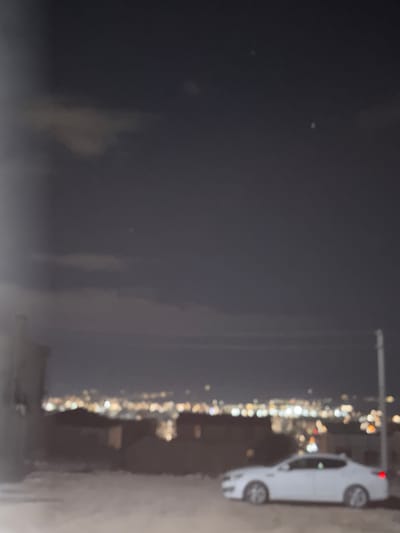 A night shot of Arinj, Armenia features a street scene with a parked white car on the right. City lights form a bright strip running vertically in the center of the frame. The sky is dark, and a blurry outline of what appears to be a telephone pole is visible near the car. The background consists of dark, undefined shapes.