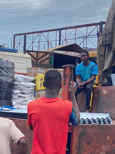 The image depicts three individuals in an outdoor setting, likely in Ogidi, Nigeria. The scene appears to be a loading area, with a truck visible in the foreground laden with goods. Two men are the primary subjects. One man, wearing a blue t-shirt with a logo and black pants, is seated on a red metallic object, possibly part of the truck's loading mechanism. He is looking towards the viewer with a neutral expression. Another man, whose back is mostly towards the camera, is wearing a bright orange t-shirt and appears to be interacting with the seated man. A third person, wearing a light-colored shirt with red trim, is partially visible in the lower right corner.
The truck is loaded with various items, including boxes wrapped in brown paper and secured with blue straps, and several white sacks. The corrugated metal roofing of a nearby structure is visible, suggesting a covered market or storage area. The overall atmosphere seems to be one of commerce or logistics. The lighting suggests it is daytime, with overcast skies. There is no discernible text in the image, apart from a logo on the blue t-shirt which is not clearly legible. The context of Ogidi, Nigeria, places this scene in a West African urban or peri-urban environment.