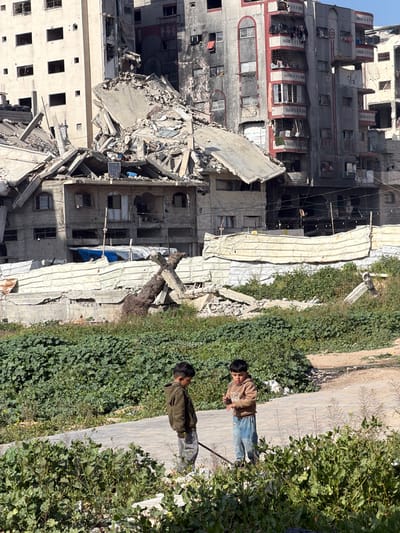 A severely damaged high-rise building stands amidst rubble. Sections are collapsed, and windows are destroyed, revealing a dark, charred interior. Two boys stand on a sidewalk next to dense green bushes. One wears a brown jacket and jeans, while the other is in a green jacket. Debris is visible on the ground near the bushes, and the building's ruins cast a long shadow over the scene. The overall impression is one of devastation and displacement.