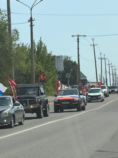 A line of cars, some decorated with Russian flags and other flags, is driving on a paved road in Kamyshin, Russia. Overhead power lines run parallel to the road. The cars are a mix of SUVs and sedans. Buildings and greenery are visible on the side of the road. A sign with Cyrillic text is also visible.