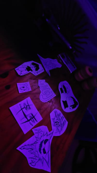 The image shows a wooden surface, possibly a table or desk, illuminated by a strong purple light source. On the surface are several hand-drawn images on pieces of paper. The subjects include cartoon characters, an eye with Japanese text within, and a ghost-like figure. Other objects present include skateboard wheels and a package labeled "CHARGERS". The lighting obscures details in the upper left corner of the image, but what can be seen suggests the presence of a skateboard. Based on the prompt, the location is Lanús Este, Argentina.