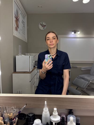 A woman is taking a selfie in what appears to be a beauty salon or medical spa. The setting includes a medical chair, beauty products on shelves visible in the mirror, and a sink area with white cabinetry. There is also a clock on the wall and a framed advertisement. While the location is suggested to be Murino, Russia, this cannot be confirmed solely from the image content.