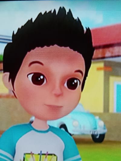 The image shows a close-up of a 3D animated cartoon character, a young boy with black spiky hair, large brown eyes, and fair skin. He is wearing a light blue and white t-shirt with white stripes on the sleeves. In the background, a light blue cartoon car is visible, along with parts of houses with red and yellow facades. The scene is set in an outdoor environment that appears to be a suburban area, possibly in Merbopuntong, Indonesia, given the architectural hints of the buildings. The lighting suggests it is daytime, with a bright sky. The overall image is of good quality with clear resolution, and the animation style is typical of children's programming.