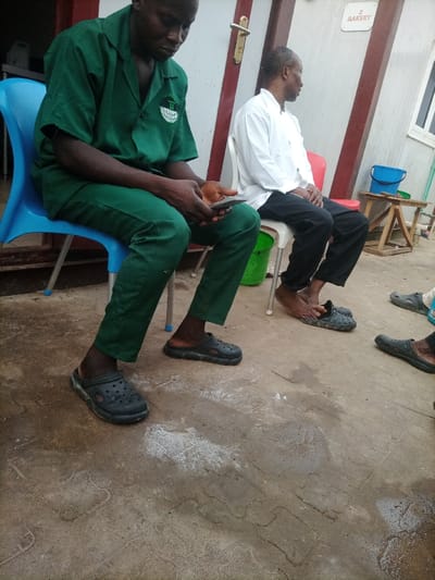 The image depicts an outdoor scene in Opete, Nigeria, featuring three dark-skinned individuals in a waiting or resting area near a building.
In the foreground, a male in a green short-sleeved uniform shirt with a white emblem on the left chest and matching green trousers is seated on a light blue plastic chair. He is looking down at a dark-colored smartphone held in both hands. He wears dark grey, perforated slip-on clogs.
To his right, a second male is seated on a white plastic chair, turned to his right and looking away from the camera. He wears a white long-sleeved shirt and dark trousers. His feet are bare, positioned above a pair of dark grey, perforated slip-on clogs. A red plastic chair is situated directly behind him. The lower legs and feet of a third individual, wearing similar dark slip-on clogs, are partially visible on the far right of the frame.
The background consists of a light-colored building with reddish-brown vertical structural elements. A white sign with "BAKERY" in red text and a numerical "2" above it is visible on the building wall. A light-colored door with a metallic handle is to the left, and a white-framed window is to the right. A green bucket is positioned under the first man's chair, and a blue bucket rests near a small wooden stool on the far right background. The ground is a patterned concrete or paved surface, exhibiting scattered white residue or stains and damp patches.