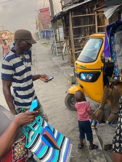 A yellow motorized tricycle sits near the curb. A young child in a red shirt leans against it. In the foreground, a man in a striped shirt and baseball cap holds a mobile phone. A colorful striped bag rests on someone's lap beside him. A building with a sign is visible in the background, along with a bicycle parked on the street. Brooms are tied to the side of the tricycle.