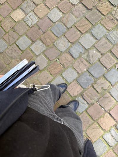 The scene is a top-down view of an individual standing on a cobblestone pavement in Ixelles - Elsene, Belgium. The ground consists of irregularly shaped, multi-colored cobblestones in shades of grey, brown, and reddish-brown, arranged in a diagonal grid pattern with some green vegetation present in the grout lines. The person is wearing dark grey jeans and black dress shoes. A dark, possibly black, outer garment covers the individual's upper legs and lower torso. The person's partially visible left hand holds a silver-colored laptop, which is partially inserted into a dark zippered protective sleeve. White wired headphones with a black zipper pull are visible, seemingly connected to or near a dark zippered bag draped over the person's left hip area. The individual appears stationary.