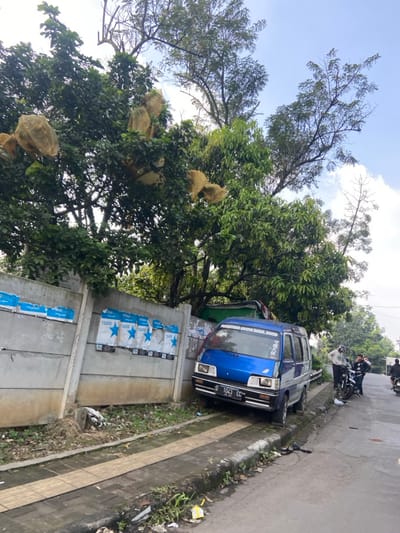 The image shows a blue minivan parked on the side of a paved road in Kecamatan Cimahi Selatan, Indonesia. The vehicle is parked facing forward with its front end closest to a concrete wall with a sidewalk between the wall and the road. There are several posters attached to the wall. The van is in a residential area and a large tree is in the foreground to the left of the vehicle, with a smaller tree to the right. The van is parked partially on the sidewalk and partially on the road. There are three people in the distance, all dressed in dark clothing, who are walking away from the van. The ground is covered in loose debris. The sky is clear and the sun is shining. The coordinates provided are -6.93 latitude and 107.565 longitude.
