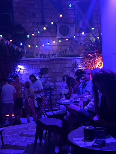 This image captures an outdoor evening scene at a venue in Herceg Novi, Montenegro. The environment is bathed in blue and purple lighting, creating a vibrant atmosphere. String lights with colorful bulbs, red and yellow bunting, and a Scottish flag are visible, suggesting a festive or themed gathering.
In the foreground, a red metal chair is prominent. To the left, a group of people are standing; one man wears a yellow t-shirt with text, and another is in a white t-shirt. Further back, more individuals are seated and standing around tables. A man in a light blue shirt is holding a cigarette and looking towards the camera. Another woman with long, dark hair is in the immediate foreground, partially obscuring the view of the man next to her. A man with long dark hair and a white t-shirt is seen in conversation with a woman.
The background features a brick wall, an air conditioning unit mounted on the wall, and lush foliage. Tables with chairs are arranged throughout the space, indicating a seating area for patrons. The overall impression is of a lively social gathering at a bar or restaurant with an outdoor patio.