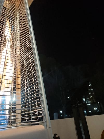 The image depicts an outdoor night scene, likely from a balcony or terrace in Tiranë, Albania, overlooking a city. In the foreground, a prominent white outdoor heater extends horizontally across the lower half of the frame. Its reflective metal grate, featuring a diamond-like mesh pattern, covers bright, glowing heating elements. These elements emit light that transitions from warm orange and yellow tones on the left to cooler blue hues on the right, suggesting the heater is actively in use.
The background is dominated by a dark night sky, indicating deep twilight or nighttime. Below the distant horizon, numerous small, scattered lights twinkle, revealing a cityscape or residential area. These lights appear as bright, unfocused points against the darkness. To the far right, a section of a light-colored, possibly white, building wall or pillar is visible, part of the structure from which the photo is taken. A dark, indistinct shape is faintly visible on this wall. No people or legible text are present. The scene conveys a calm, cool evening, with the heater providing warmth in an elevated outdoor setting overlooking the illuminated urban landscape.