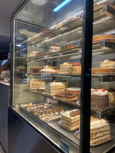 This is an eye-level, slightly downward-angled shot of a refrigerated display case filled with an assortment of cakes and pastries, likely in a bakery, cafe, or patisserie in San Fernando del Valle de Catamarca, Argentina. The image is oriented vertically, showing the contents across multiple shelves.
The display case is brightly lit by LED strips lining the shelves, creating prominent reflections on the clear glass front. These reflections show overhead lighting, parts of the ceiling, and some interior elements of the establishment. A bright, cool-toned fluorescent light fixture is visible on the left side, reflecting off the glass.
The shelves are laden with numerous cakes of various types. Visible options include layered cakes with cream and fruit toppings (like cherries), chocolate-covered cakes, cakes with intricate patterns of nuts or crushed biscuits, roll cakes, and some tart-like pastries with chocolate designs. Many cakes are presented as individual slices or smaller portions, while others appear to be whole cakes. The textures range from smooth cream and chocolate glazes to crumbly toppings and layered sponges.
Visible text includes the reflection of the word "Coffee" along with a coffee cup icon, and partially legible text that appears to be "enjoy your life" or similar, suggesting the establishment also serves coffee. On a lower shelf inside the case, a label or sign is visible with what appears to be "San Cruz" or "San Cru..." accompanied by a snowflake or star logo, and a phone number: "2144-3886".
The scene captures a static moment, showcasing the baked goods available for purchase, emphasizing their variety and appealing presentation.