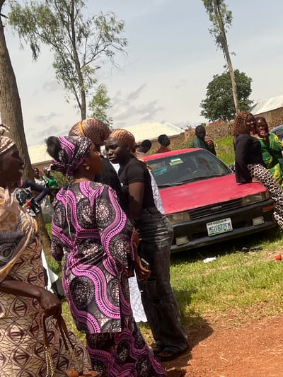 In an outdoor setting in the city of Unknown, Nigeria, a group of people are gathered around a red Toyota sedan. The sky is partly cloudy, and trees with sparse foliage are visible in the background. The ground is a mix of grass and reddish-brown dirt.
Several individuals are the focus of the image. In the foreground, a woman wearing a light-colored, patterned dress and a headscarf is holding a brown purse with a gold chain. To her right, another woman is wearing a vibrant pink and black patterned dress and a matching headscarf. Her gaze is directed towards a young woman in a black t-shirt and distressed denim jeans, who appears to be speaking to her. This young woman has blonde-streaked hair.
Further back, a man in a green shirt is standing near the car, holding a red object. Other individuals are visible in the background, including a woman in a green outfit with a patterned skirt, and several men. The red Toyota has a license plate that reads "MGU 672FM PLATEAU". The car appears to be parked on the dirt and grass area. The overall scene suggests a social gathering or event.