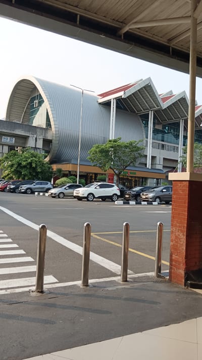 The image shows an exterior view of the Soekarno-Hatta International Airport's Terminal 3 in Tangerang, Indonesia. The distinctive curved, metallic roof of the terminal building dominates the background, with its modern architectural design featuring segmented, ribbed sections. Several cars are parked in a lot in front of the terminal, which has glass windows and a series of peaked, red-roofed sections extending from the main structure. Trees and some greenery are visible between the parking lot and the building. In the foreground, a pedestrian crossing with white stripes is marked on the asphalt road, with several polished metal bollards positioned along its edge. The sky is clear and appears to be daylight. The scene is static and depicts a typical airport environment.