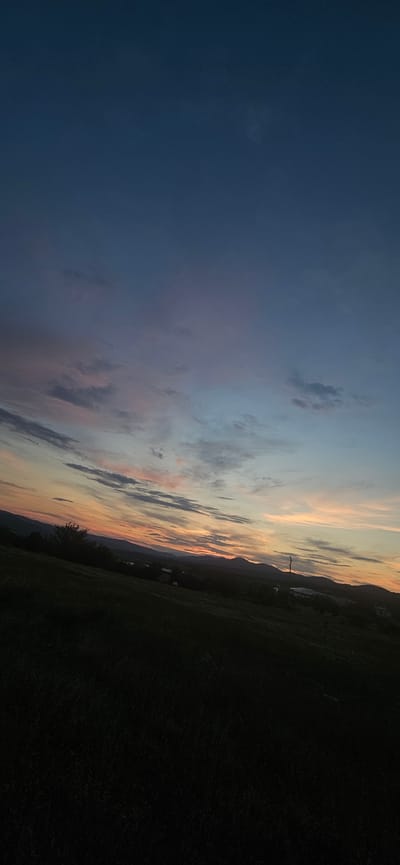 This image depicts a rural landscape at dusk, featuring a rolling hill in the foreground covered in dark vegetation, possibly grass or low-lying crops. The midground reveals a silhouette of distant mountains, with a few faint structures and trees visible against the horizon. The sky dominates the upper portion of the image, transitioning from a deep blue at the top to hues of pink, orange, and light blue near the horizon, indicative of a sunset or twilight. Wispy clouds are scattered across the sky, catching the fading light. The scene appears to be located in Momin Prohod, Bulgaria, given the presence of rolling hills and mountainous terrain characteristic of the region. There are no people or animals visible in the image, nor are any specific actions or interactions taking place. The overall mood is serene and tranquil, capturing the end of the day in a natural setting.