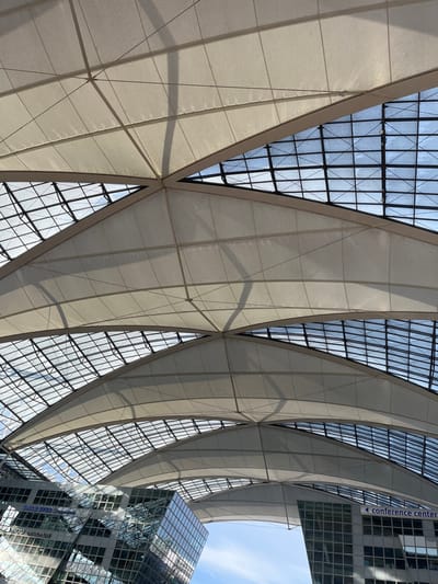 The image displays a forensic view of the Munich Airport Center (MAC Forum) at Munich Airport, located in Oberding, Germany. The scene is dominated by a vast, vaulted tensile membrane roof system, composed of light-colored, semi-translucent fabric panels structurally supported by a network of dark beams. Interspersed within the fabric are extensive glass skylight sections, organized in a grid pattern, through which a clear blue sky is visible. Shadows from the roof's internal structure are cast across the fabric surfaces. Below the roof on the left, a modern, multi-story building features a reflective glass facade that mirrors the sky and portions of the roof. On the right, another building with a glass facade is visible, displaying a dark blue sign with a white left-pointing arrow and the text "conference center." A segment of clear blue sky is visible between these two structures, beneath the roof arch. No individuals, vehicles, or specific actions are depicted within the frame.