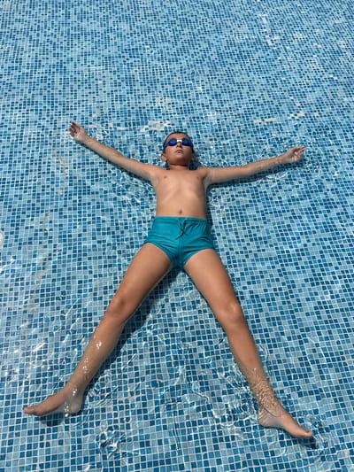 A young person with brown hair and blue swimming goggles lies on their back in a swimming pool, arms and legs spread wide, resembling a star shape. The person is wearing blue swim trunks and is submerged in clear water, with their body partially visible beneath the surface. The bottom of the pool is tiled with small blue and white squares, creating a mosaic pattern that is distorted by the ripples on the water's surface. Sunlight filters through the water, casting shimmering patterns and highlighting the contours of the person's body. The scene appears to be a sunny day, likely during the summer, given the water activity. The person's pose suggests relaxation and enjoyment, possibly floating or enjoying a moment of rest in the pool. The setting is an outdoor swimming pool, indicated by the tiled bottom and the natural light. No visible text is present in the image. The location context of Grivitsa, Bulgaria, places this scene within that geographical region.