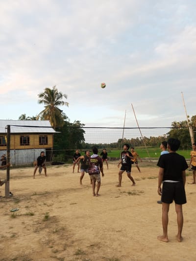 A group of young men are playing volleyball on a dirt court in Turumame, Indonesia. The court is surrounded by trees and vegetation, with a traditional wooden house visible in the background. The sky is overcast with a few clouds. A volleyball is in mid-air, about to be hit by one of the players. The players are wearing casual clothing, and most are barefoot. The scene appears to be a casual game among friends.