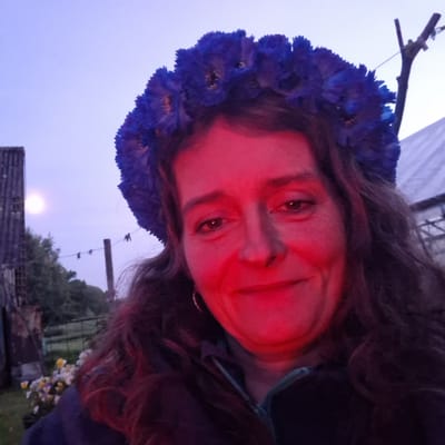 A person is shown from the chest up, wearing a flower crown made of blue flowers and adorned with dark, long hair. The image is a close-up selfie with a red light cast on the person's face, obscuring some details. The person has light-colored eyes and appears to be smiling. The background shows a twilight sky with a full moon visible. There are also hints of trees and some structures in the distance, suggesting an outdoor setting. The overall mood is somewhat mysterious due to the red lighting and twilight. The image appears to be taken at dusk. There is no visible text.