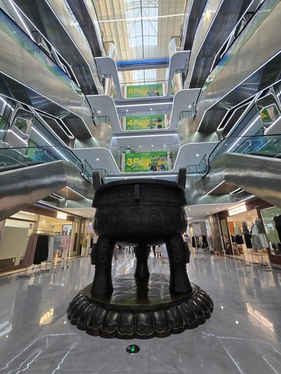 The image depicts the interior of a modern, multi-story shopping mall, likely located in Shangcheng District, China, as suggested by the context. A large, ornate bronze cauldron, possibly a decorative centerpiece, dominates the foreground. The cauldron rests on a base resembling lotus petals and features intricate carvings.
Behind the cauldron, a central atrium rises through multiple levels, characterized by sleek, metallic escalators and walkways. The architecture is contemporary, with clean lines and polished surfaces that reflect light, enhanced by linear white LED lighting accentuating the edges of the escalators and walkways. Suspended banners with Chinese text and graphics are visible on different levels, advertising various floors or promotions.
The lower levels of the mall reveal storefronts, with some displaying clothing on racks and mannequins. The floor is a polished, reflective gray marble or tile, with faint white lines. A green directional arrow icon is visible on the floor in the lower right quadrant, indicating a pathway. Several individuals are present, walking in the background, though their faces are not clearly discernible. The overall impression is one of a bustling, contemporary retail environment with an artistic, traditional element.