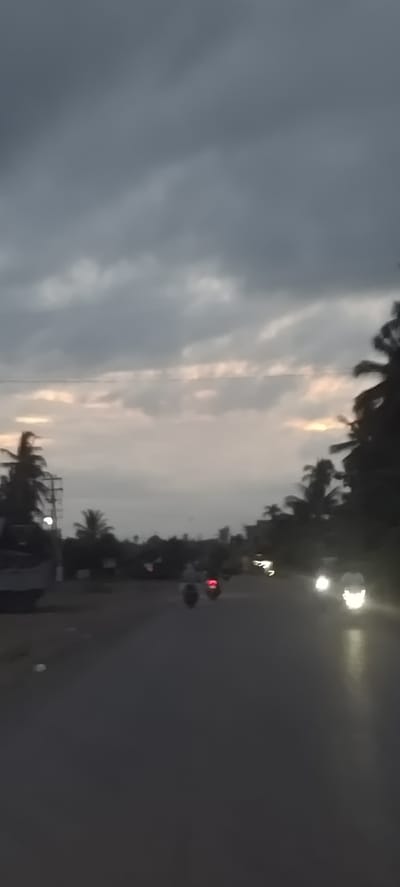This image captures a road scene in Ujongreuba, Indonesia, during twilight, likely dusk or dawn, under a heavily overcast sky. The sky is dominated by dark grey clouds, with faint, lighter patches visible along the horizon, suggesting the sun's position either below or heavily obscured by the clouds.
The foreground features a dark, paved road, appearing wet or reflecting light due to the low light conditions. Several vehicles are visible, indicated primarily by their active lights. In the mid-ground, two motorcycles can be seen, one displaying a bright red taillight. Further down the road on the right, multiple bright headlights are visible, belonging to approaching vehicles, possibly more motorcycles or cars.
Along both sides of the road, lush tropical vegetation, including numerous palm trees, forms dark silhouettes against the dim backdrop. On the left side of the road, a utility pole with overhead wires is discernible amidst the foliage. The overall lighting is low, resulting in a somewhat blurred appearance, especially for the moving vehicles. No text is visible in the image.
