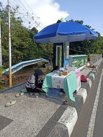 This outdoor scene, set in Mataram, Indonesia, depicts a small vendor stall operating on a bright, sunny day. The clear sky with scattered clouds suggests good weather. Lush green trees and vegetation form the backdrop, with a distinctive blue pipeline structure visible further back.
A prominent blue umbrella provides shade over a small, portable table. The table is adorned with two tablecloths: a white one featuring the word "LOVE" and a light green, patterned one. Displayed on the table are several clear jars filled with snacks, such as biscuits or nuts, along with a small blue sign that likely lists products or prices.
One woman, wearing a black hijab and a black-and-white patterned long-sleeved shirt, sits on the ground beside the table. She is leaning on her hand, looking down, appearing calm or pensive, possibly resting or awaiting customers. Nearby are a pair of sandals, a green tray, and a beverage in a plastic cup. A second individual is partially visible behind the table.
The stall is positioned on a textured paved surface adjacent to a road, which features concrete curbs and a white painted line. Further along the road to the right, piles of coconuts are visible on the ground, suggesting the presence of other vendors or goods for sale. In the background, a dark green structure and utility poles are also present. A pink sign near the table has partially visible text, including "Maso...".