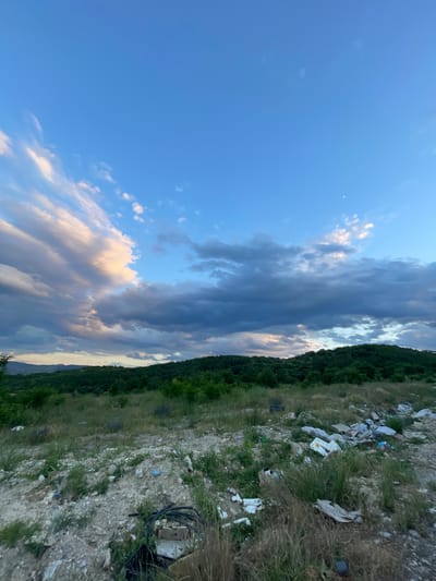 This is a photograph of an area in Ablanitsa, Bulgaria, likely a roadside or undeveloped lot. The foreground is littered with various types of refuse, including plastic bags, cardboard boxes, and other debris, scattered amongst dry grass and rocks. In the middle ground, a dense green forest covers rolling hills. The background features a vast blue sky with scattered clouds, some of which are illuminated by the setting or rising sun. A crescent moon is visible in the upper right portion of the sky. There are no people or animals depicted in the image.