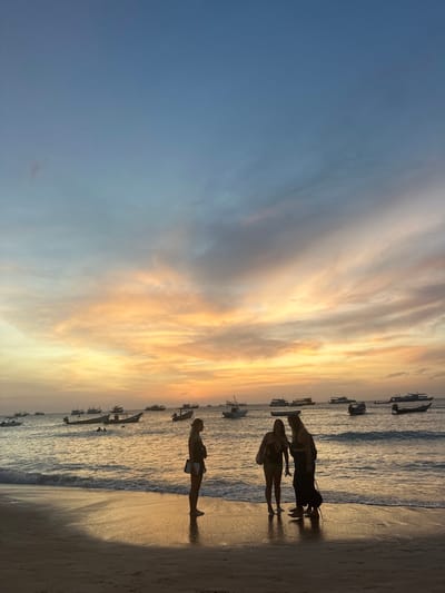 The image captures a sunset over a beach in Ko Tao, Thailand. The sky displays a gradient from deep blue at the top, transitioning to light blue, and then to a broad band of orange, yellow, and pink hues with scattered clouds near the horizon. The calm ocean water, reflecting the warm sunset colors, occupies the midground. Numerous boats, primarily traditional Thai longtail boats and other small vessels, are visible as dark silhouettes against the illuminated water and sky. The foreground shows a wet sandy beach, reflecting the ambient light. Three individuals are positioned on the wet sand near the waterline, entirely silhouetted. The individual on the left stands facing slightly right, holding a bag. The middle and right individuals are facing each other, engaged in an apparent interaction or conversation. Two additional human figures are partially visible in the water further from the shore.