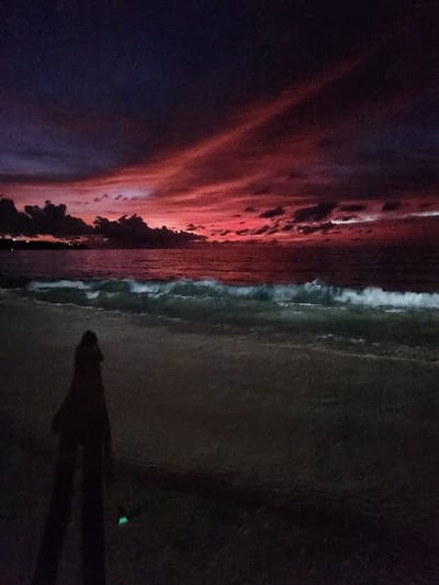 The image depicts a beach scene at dusk in Pecatu, Indonesia. The sky transitions from dark purple and blue overhead to vibrant reds and oranges along the horizon, marked by dark, scattered clouds and two prominent diagonal red streaks in the upper atmosphere. The ocean, appearing dark, exhibits white-capped waves breaking and rolling towards the shore. A distant, silhouetted landmass with faint lights is visible on the left horizon. In the lower-left foreground, a solitary human figure stands in silhouette on the dark sand, facing the water. A small, bright green glowing object is present on the sand near the figure's feet.