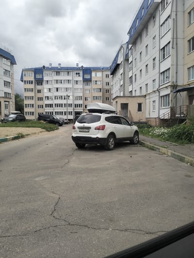 The image depicts an outdoor urban scene on an overcast day. In the foreground, a white Nissan Qashqai SUV with a roof box is parked on a cracked asphalt road. The car's license plate reads "H048YM 199". To the left of the SUV, a black SUV and a black sedan, also with roof boxes, are parked on a patch of grass next to a sandy area.
In the background, multi-story apartment buildings with blue-tiled roofs dominate the skyline. The buildings appear to be residential, with numerous windows visible on their facades. One building on the right has external staircases leading to entrances. The environment appears to be a residential courtyard. The scene is devoid of any visible people. The location is identified as Zyuzino, Russia.