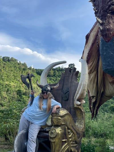 A woman, dressed as a Viking with a helmet and holding an axe, poses on a large, ornate throne-like sculpture. The throne is decorated with large horns and metallic gold accents. The woman is wearing a light blue t-shirt and grey sweatpants. Behind her, a large dragon statue is partially visible, with its wing and head in the frame. The scene is set outdoors, with lush green hills and a blue sky with white clouds in the background. This location is identified as Thôn 1, Vietnam.