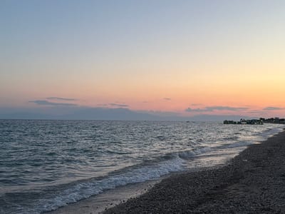 A tranquil scene unfolds on a pebble beach at sunset in Nea Flogita, Greece. The sky above is a gradient of soft blues and warm oranges, transitioning from a pale azure at the zenith to a soft peach and pink hue near the horizon. Wisps of clouds are scattered across the sky, catching the last rays of the sun.
The calm, dark blue sea laps gently at the shore, with small waves creating a foamy white edge as they meet the pebbles. The beach itself is covered in dark, smooth pebbles. In the distance, silhouetted against the colorful sky, are mountains, hinting at the mainland or a nearby island.
Further along the coastline, there are hints of human activity. A few small boats are visible on the water, and near the right edge of the frame, there appears to be a floating water park with inflatable structures. Lights are also visible on the shore in the far distance, suggesting a populated area or resort. The overall atmosphere is peaceful and serene, evoking a sense of calm and relaxation as the day draws to a close. No text is visible in the image.
