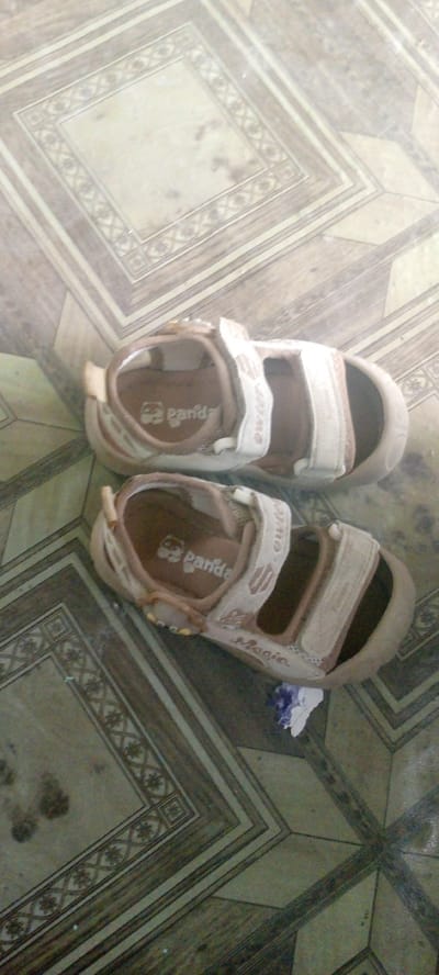 Two children's sandals are lying on a floor with a patterned tile design. The sandals are beige and white, with velcro straps. The sandal on top has a logo with "panda" written on it, and the sandal on the bottom has "Magic" written on it. A small piece of purple and white paper is partially visible under the sandal on the bottom. The scene appears to be indoors, possibly in a home, in Marhamat, Uzbekistan.
