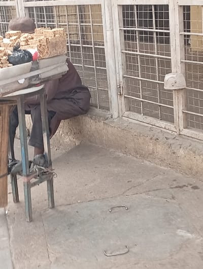 In Gawuna, Nigeria, an outdoor scene depicts a street vendor operating near a building. A person, partially visible and seated, wears a dark brown garment and a brown cap, with their feet resting on the lower crossbar of a simple vendor's table. The table features an unpainted wooden top supported by light blue/grey painted metal legs. On the table, multiple stacks of light brown, rectangular items (likely food products) are displayed for sale. A black plastic bag and a small red object are also on the table. Behind the vendor, a building facade with white or light-colored metal grates covers window or door openings, forming a grid pattern. A raised concrete ledge runs along the base of this structure. Attached to the right-side vertical grate frame is a white, helmet-like object with two dark circular markings. The ground is a light grey concrete surface with minor imperfections. Two U-shaped metal handles are embedded in the concrete in the foreground. The scene is illuminated by natural light.
