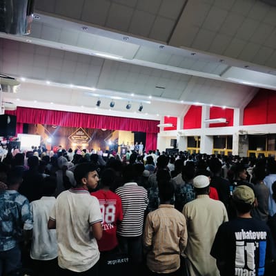 A large crowd is gathered in an auditorium-style venue in Lalmohan, Bangladesh. A stage is visible at the front, with a red curtain backdrop and an elaborate screen or backdrop with an inscription. A group of people stand on the stage. The crowd, mostly men, are facing the stage. The ceiling of the hall is high, with rows of lights.