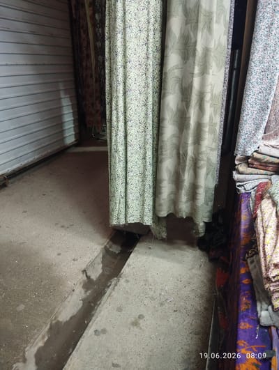 The image shows a narrow alleyway with a concrete floor. On the left, there is a closed metal shutter. In the center, several pieces of fabric are hanging, some with floral patterns and one with a leaf-like pattern. To the right, a stack of fabric rolls is visible. A drainage ditch runs along the center of the alleyway. The overall scene appears to be an outdoor market stall in the city of Istaravshan, Tajikistan, given the timestamp on the image. There are no people or animals visible in the image. The lighting suggests it is daytime.