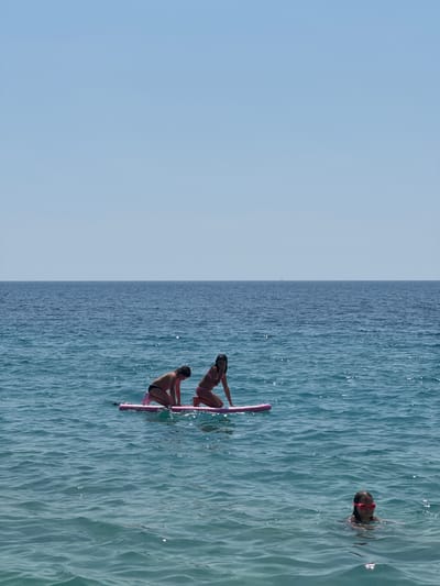 The image shows a wide, horizontal shot of the sea under a clear blue sky. The water is a vibrant blue, with gentle waves reflecting the sunlight, creating a shimmering effect. On the right side of the frame, three people are visible in the water. One person, wearing pink goggles, is swimming with their head above water. Further to the left, two people are sitting on a pink paddleboard. The person in front is leaning back on their hands, while the person behind is crouching. All three individuals appear to be enjoying a recreational activity on a sunny day, likely in the afternoon given the bright light and clear sky. The scene suggests a peaceful and leisurely day by the sea in a location with calm waters. The background is dominated by the expanse of the sea and sky, with no visible landforms or structures, emphasizing the open water setting.