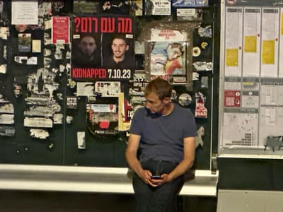 A male individual is seated on a light-colored bench at a bus stop in West Jerusalem, Israel, during nighttime. He possesses short, light brown hair and wears a dark blue t-shirt and dark pants. The man holds a dark-colored smartphone with both hands, his gaze directed downwards at the screen. Behind him and to his left, a dark wall is densely covered with numerous overlapping and torn posters and flyers, primarily in Hebrew. A prominent poster prominently displays two male portraits, with the English text 'KIDNAPPED 7.10.23' in red and black, and Hebrew text above that translates to 'What about Rom?'. Other posters feature various graphics and Hebrew text, with some partial English visible, including 'EN[D] THE FUCKING WAR'. To the man's right, a vertical white and yellow information panel, likely a bus schedule display, features multiple columns of printed information under numbers such as 22, 19, and 17. The scene is illuminated by artificial light sources, casting reflections on the surfaces.